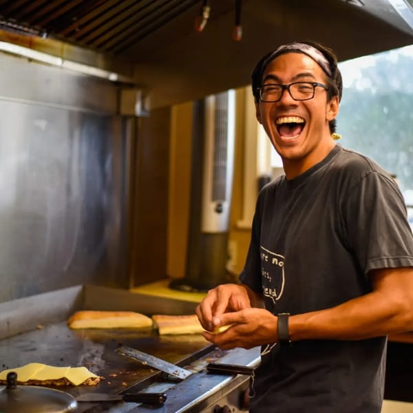 A smiling chef with glasses and a headband cooks food on a flat-top grill at Gem Grill & Pizzeria.
