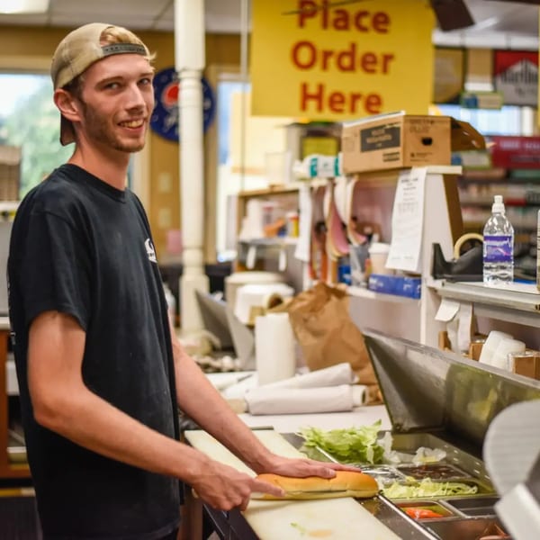 A young man in a baseball cap prepares a sub sandwich at Gem Grill & Pizzeria.